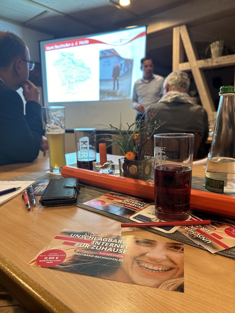 Informationsveranstaltung zum Glasfaserausbau in Bechhofen: Teilnehmer sitzen bei Getränken an einem Tisch, während ein Referent vor einer Projektionsleinwand über den Ausbau informiert; im Vordergrund Flyer mit dem Slogan ‚Unschlagbares Internet für Zuhause‘.