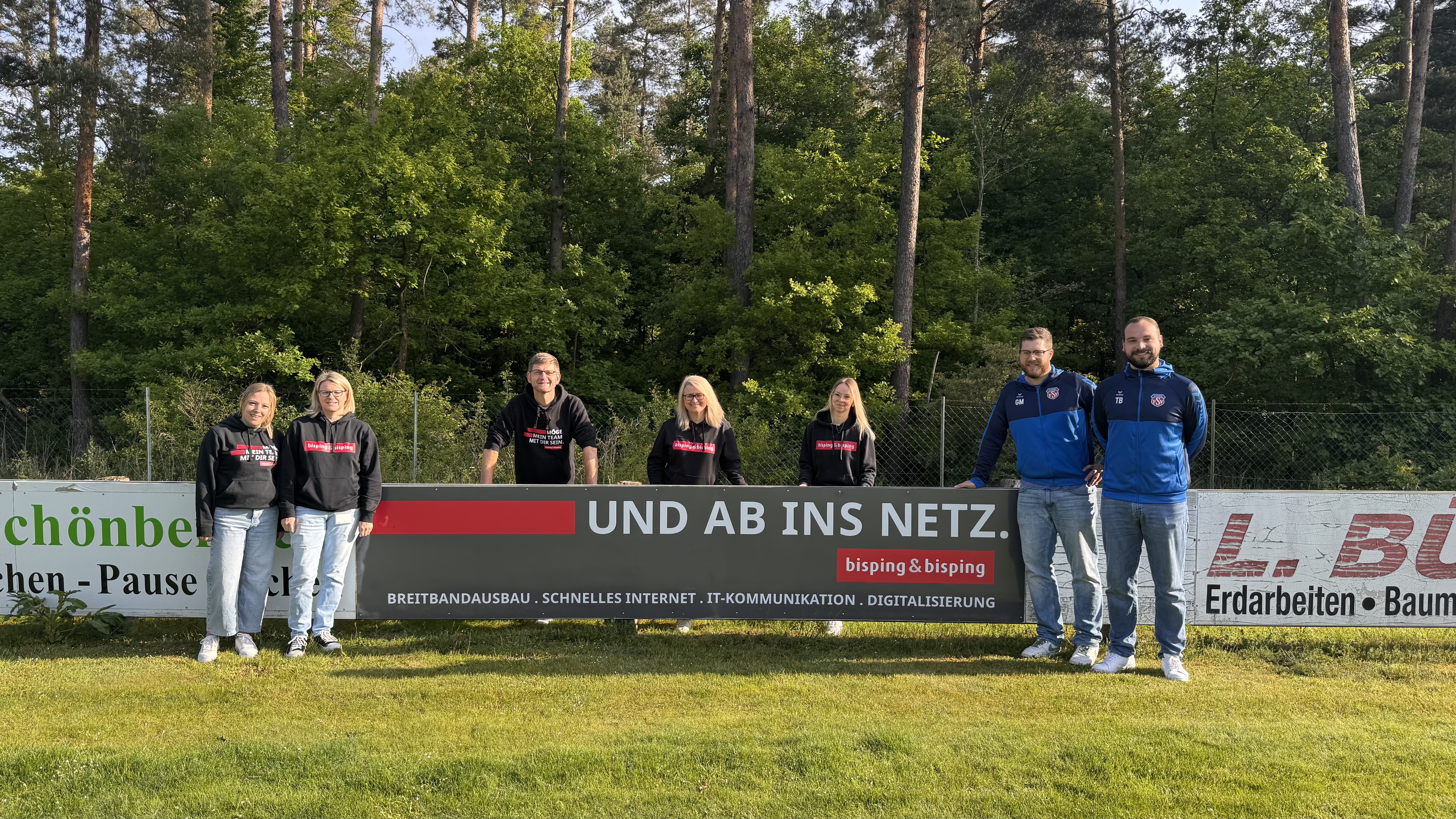 Team von Bisping & Bisping und Vertreter des Sportvereins Schönberg präsentieren gemeinsam eine neue Werbebande mit der Aufschrift ‚Und ab ins Netz‘ – symbolisch für die Partnerschaft im lokalen Vereinsumfeld.